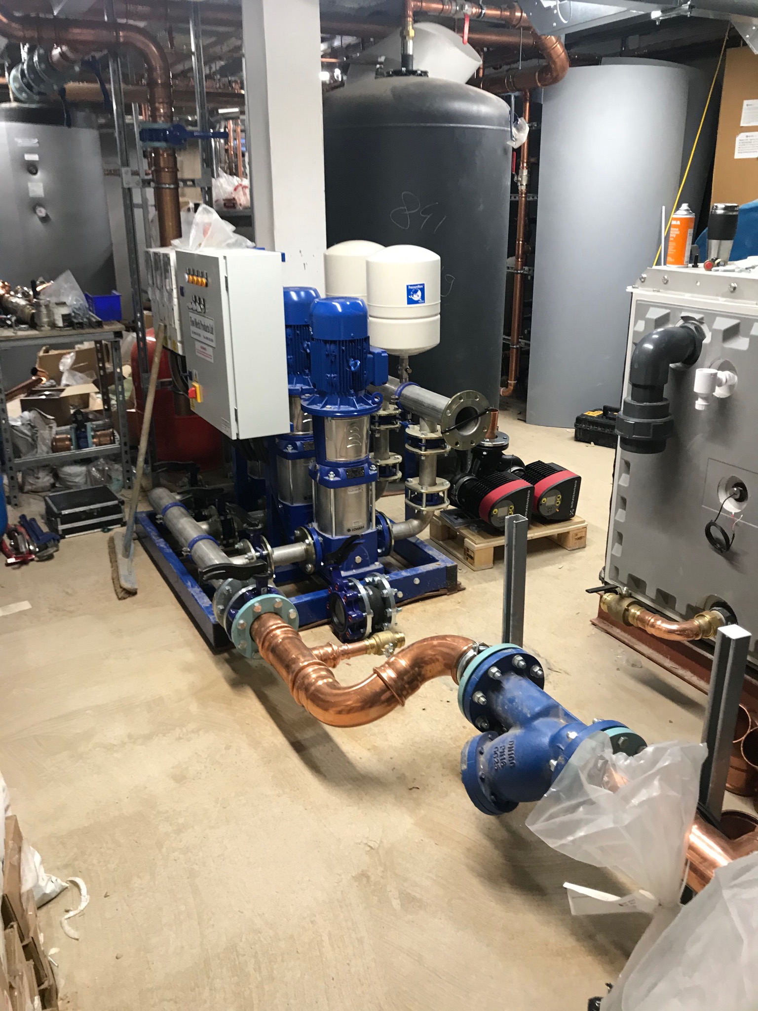 Instalacja pompowa z miedzianymi rurami i niebieskimi zaworami w pomieszczeniu technicznym, widoczne zbiorniki ciśnieniowe i szafa sterownicza, w tle regał z narzędziami i akcesoriami.