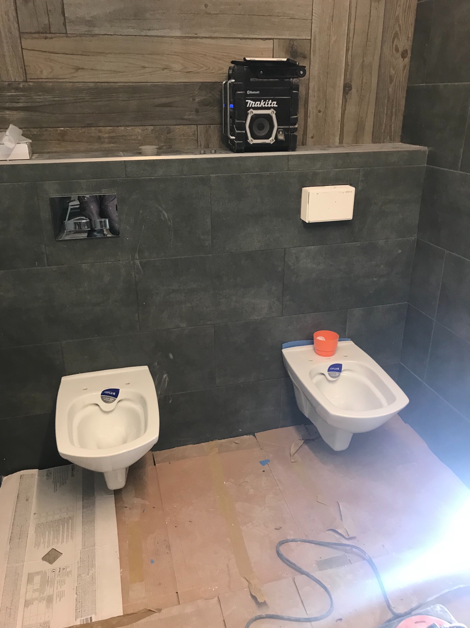 Montaż dwóch białych bidetów Cersanit w łazience z szarymi płytkami, widoczny głośnik Makita na półce i fragment podłogi w trakcie prac wykończeniowych.