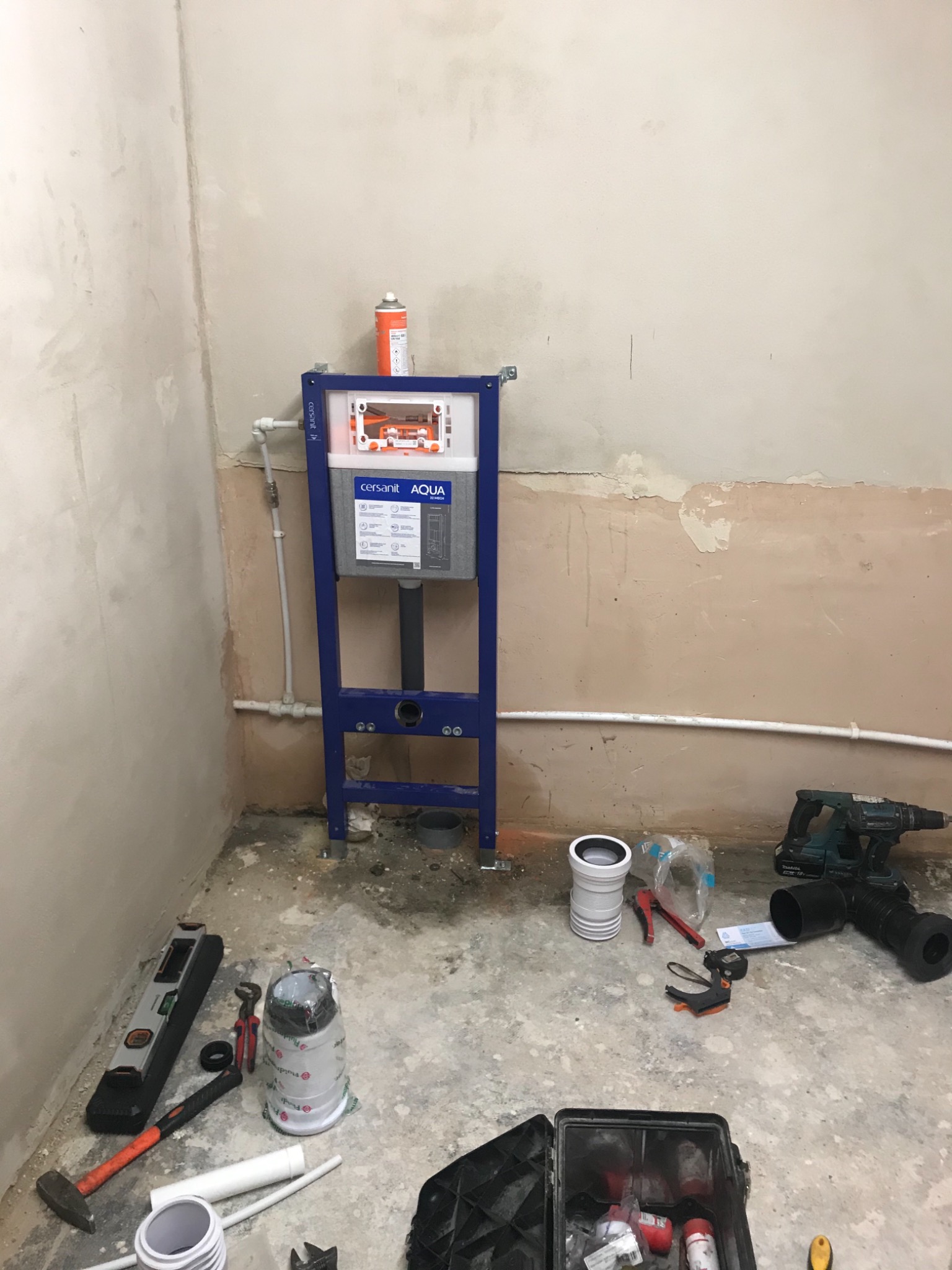 Montaż stelaża podtynkowego Cersanit Aqua w surowym wnętrzu łazienki, widoczne narzędzia hydrauliczne, poziomica, wiertarka akumulatorowa Makita, puszka pianki montażowej. Ściany przygotowane...