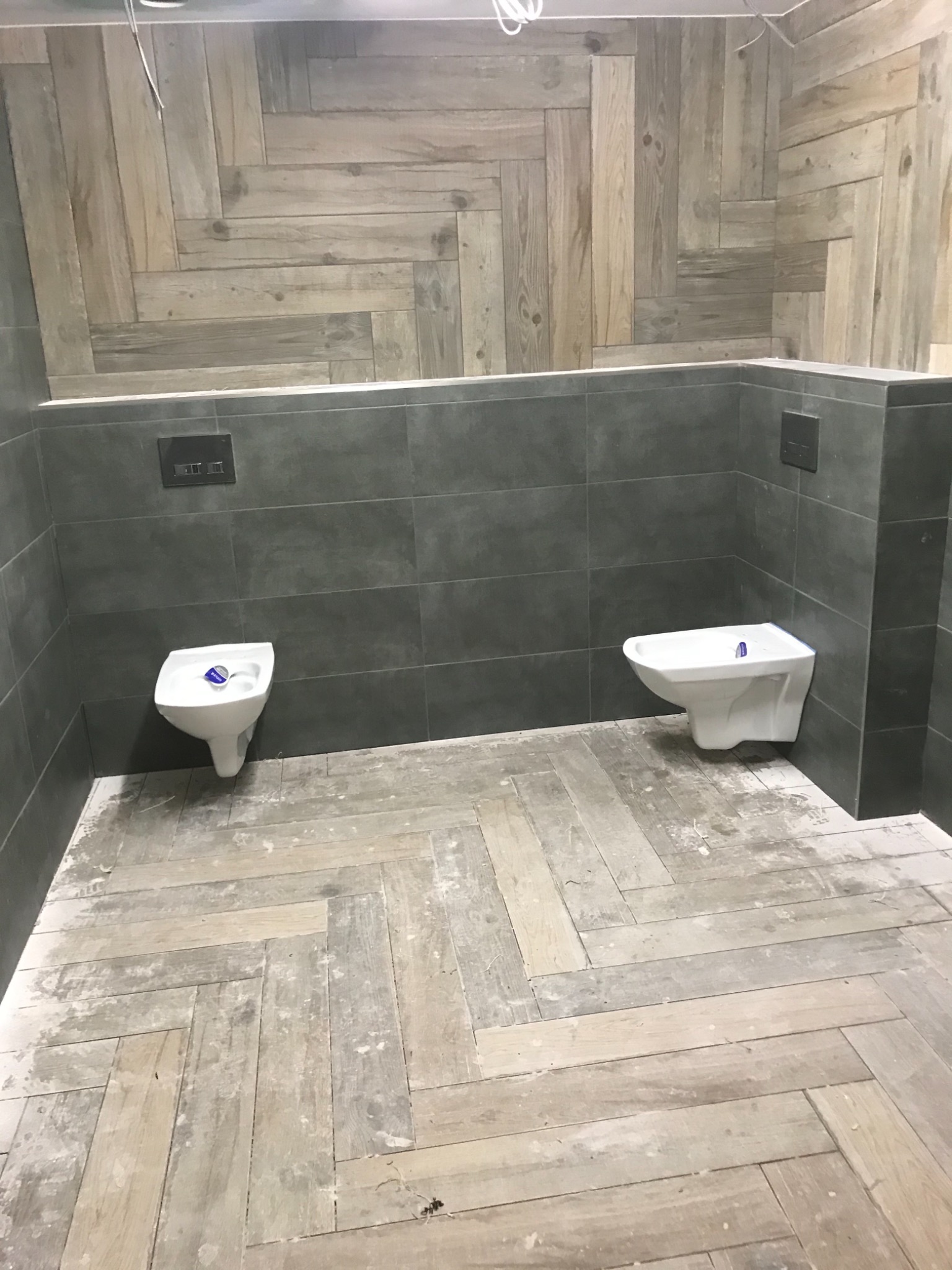 Dwa białe bidety zamontowane na szarej, kafelkowej ścianie, podtynkowy system spłukiwania widoczny powyżej, podłoga w drewnopodobną jodełkę, ślady po remoncie.