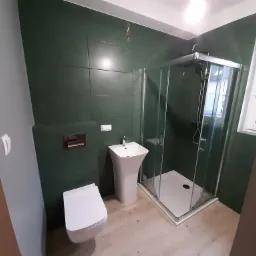 Nowoczesna łazienka z zielonymi płytkami, kabiną prysznicową z transparentnego szkła, wiszącą toaletą i umywalką na postumencie, oświetlona światłem dziennym z okna.