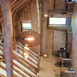 Wnętrze rustykalnego pomieszczenia z drewnianą konstrukcją dachu, kamiennymi ścianami, terakotową podłogą ułożoną w jodełkę oraz dekoracyjnymi lampami wiszącymi z żarówkami Edisona.