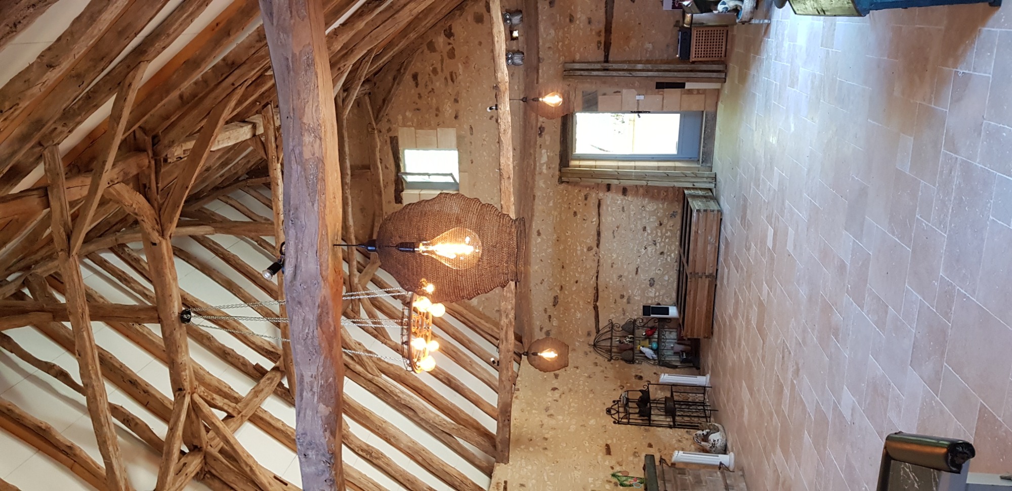 Wnętrze rustykalnego pomieszczenia z drewnianą konstrukcją dachu, kamiennymi ścianami, terakotową podłogą ułożoną w jodełkę oraz dekoracyjnymi lampami wiszącymi z żarówkami Edisona.