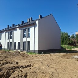 Usługi ogólnobudowlane Błażej Wojtaczka - Nowoczesne, szeregowe domy w trakcie budowy, z białymi elewacjami i szarymi akcentami, na tle błękitnego nieba. Wykopana ziemia przed budynkami.