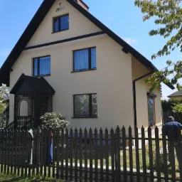 Jasny, dwukondygnacyjny dom z ciemnym dachem i stolarką okienną, wykończony tynkiem, z datą '1997' widoczną na szczycie, ogrodzony ciemnym płotem, z fragmentem pracownika w niebieskiej koszuli...