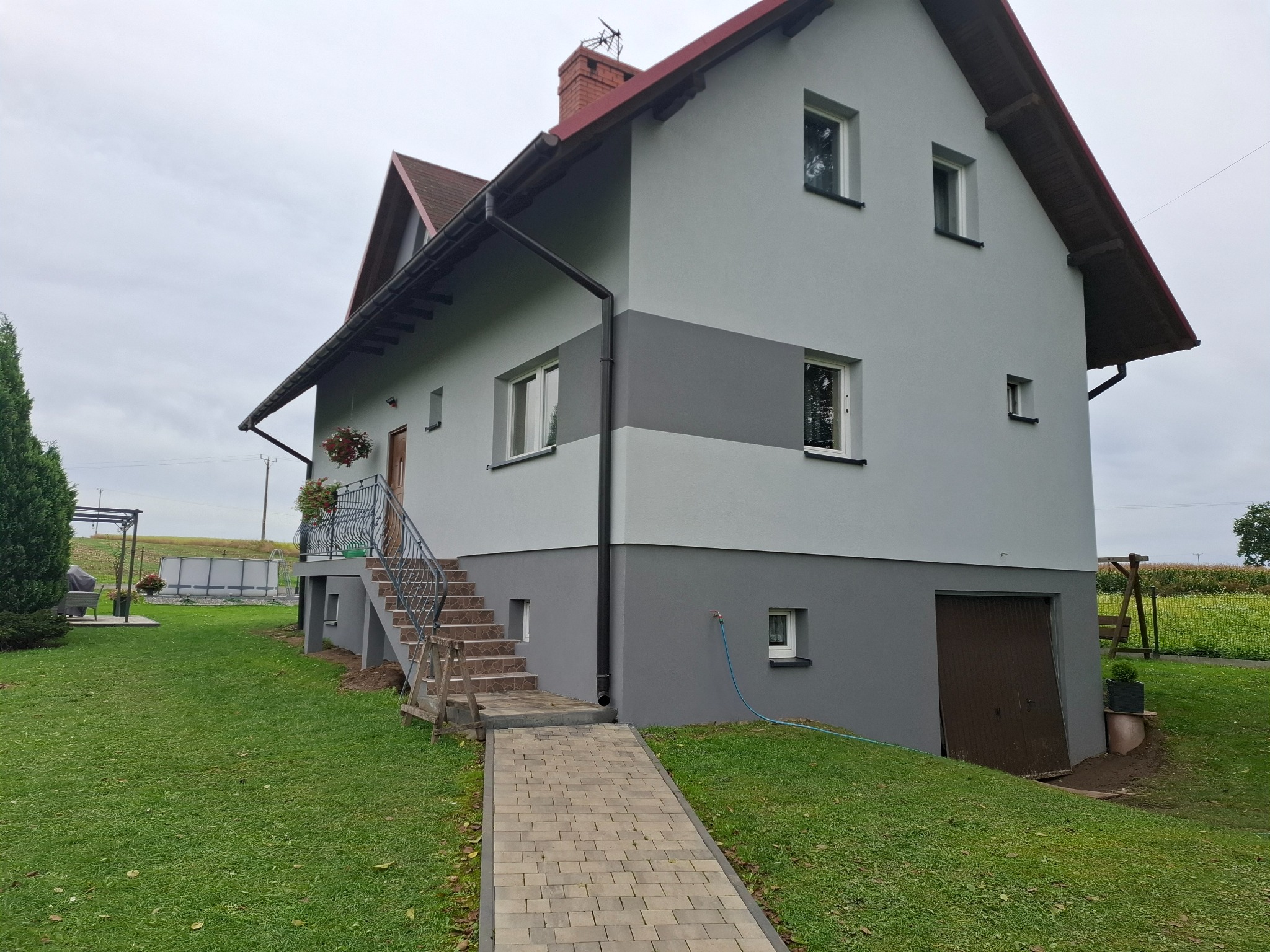 Dwukondygnacyjny dom z szarą elewacją, ciemniejszym pasem na środku i brązowym dachem, widziany z perspektywy ogrodu z kostką brukową.