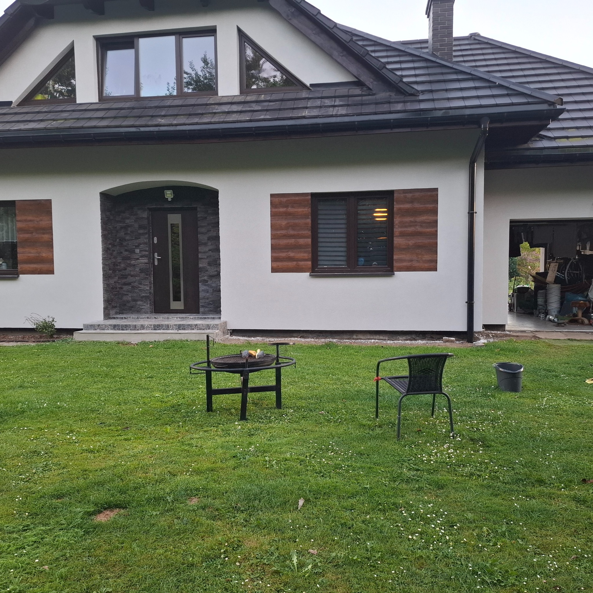 Elewacja domu jednorodzinnego z elementami dekoracyjnymi imitującymi drewno wokół okien i kamień przy wejściu. Widoczny trawnik z grillem i krzesłem.