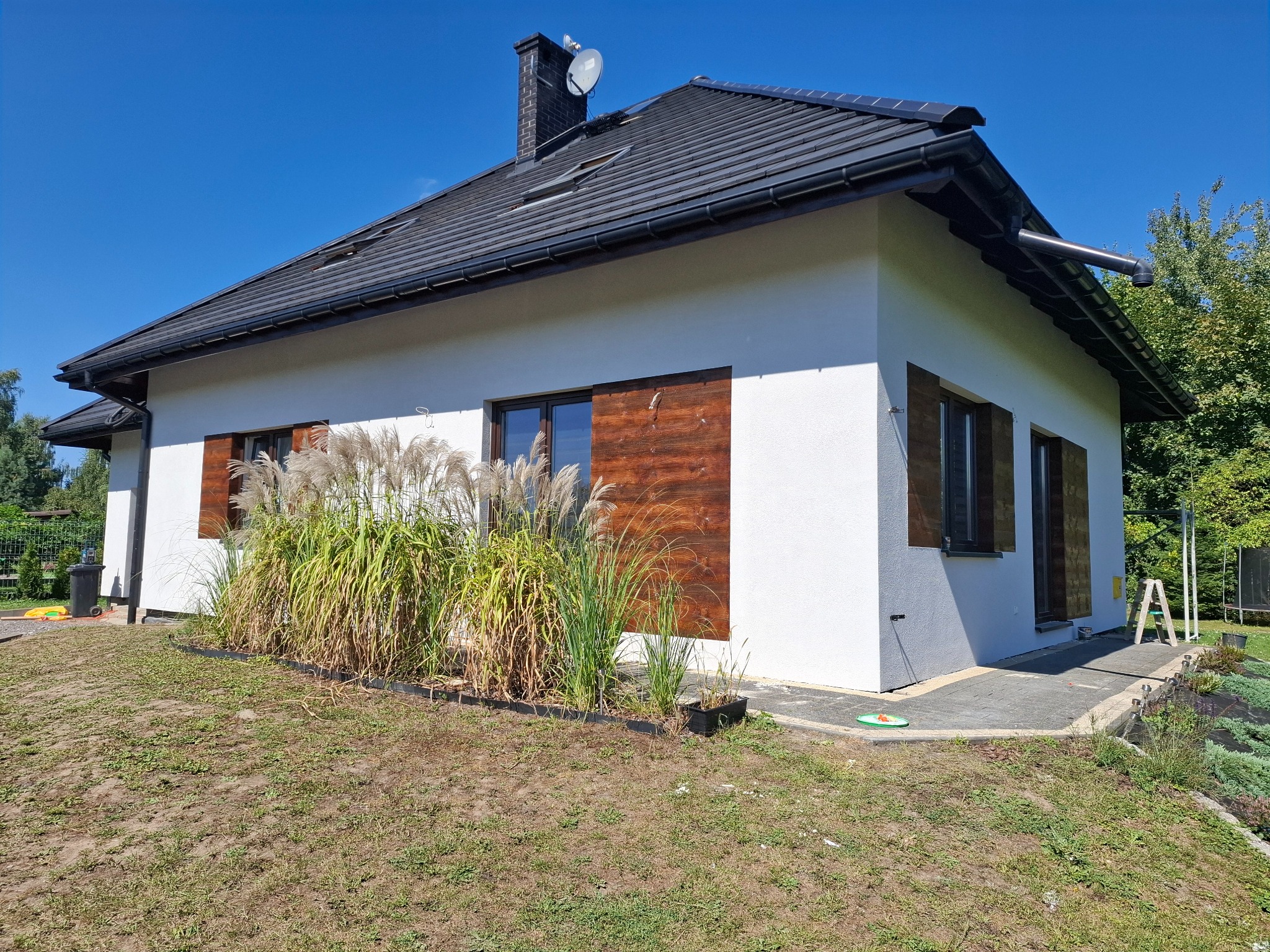 Biały dom jednorodzinny z ciemną dachówką i drewnianymi okiennicami, otoczony zielenią. Widoczne detale wykończenia elewacji i zagospodarowania terenu.