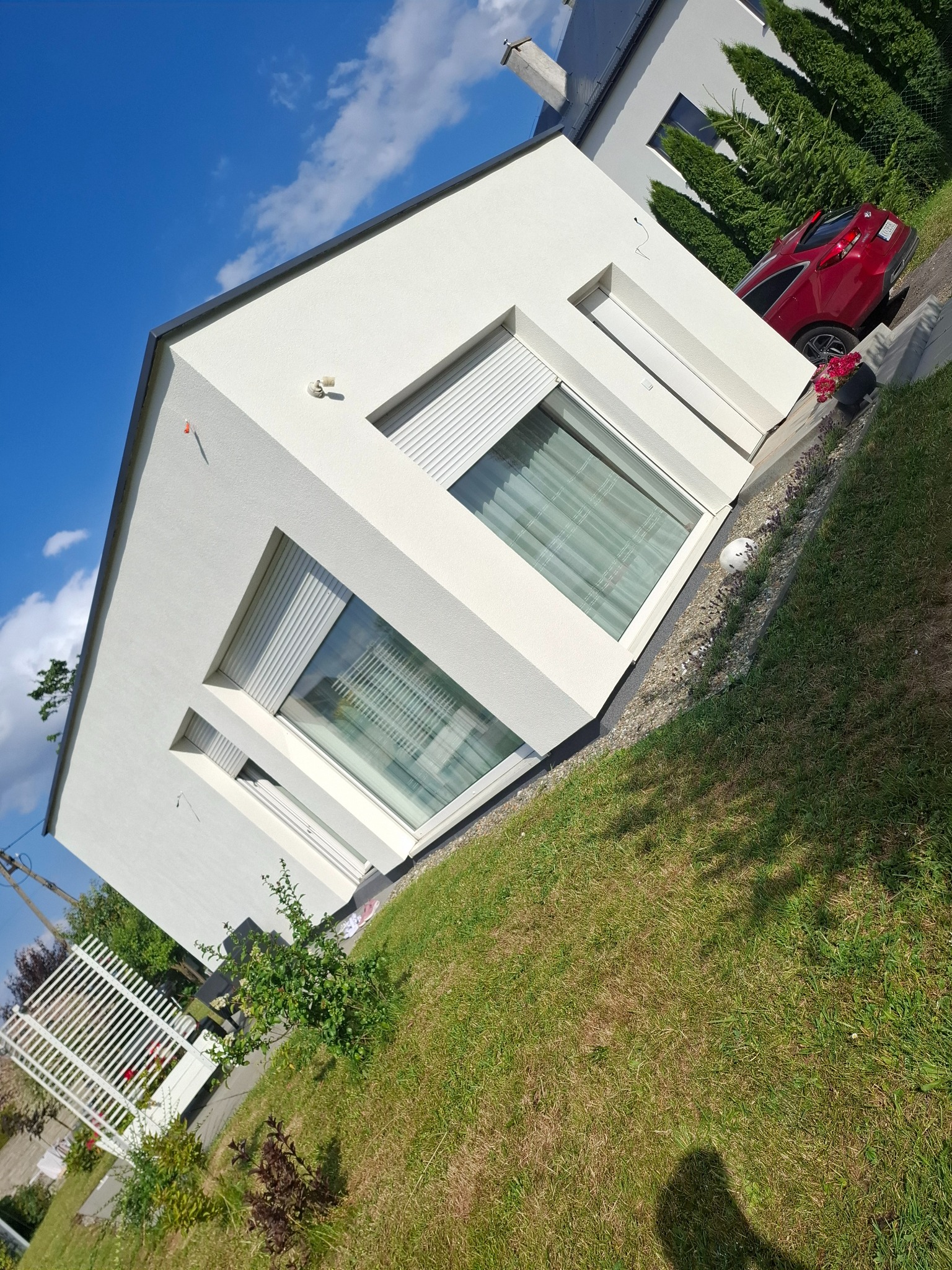 Nowoczesny, biały dom jednorodzinny z płaskim dachem, widoczny z boku, z roletami zewnętrznymi i zadbanym trawnikiem. Czerwony samochód w tle. Słoneczny dzień.