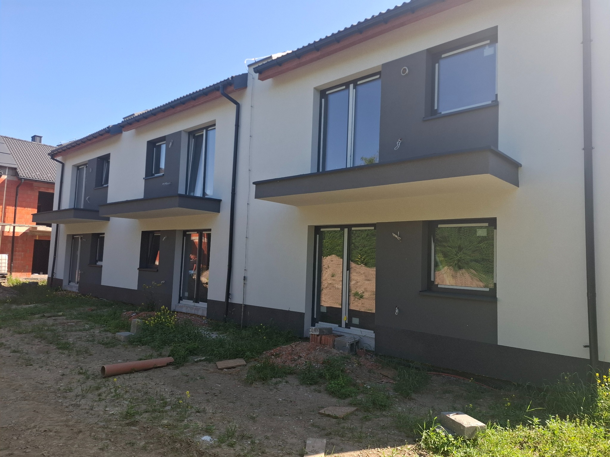 Nowoczesne, dwukondygnacyjne domy w zabudowie szeregowej z balkonami, wykończone w bieli i szarości, w słoneczny dzień. Widoczne elementy wykończeniowe elewacji.