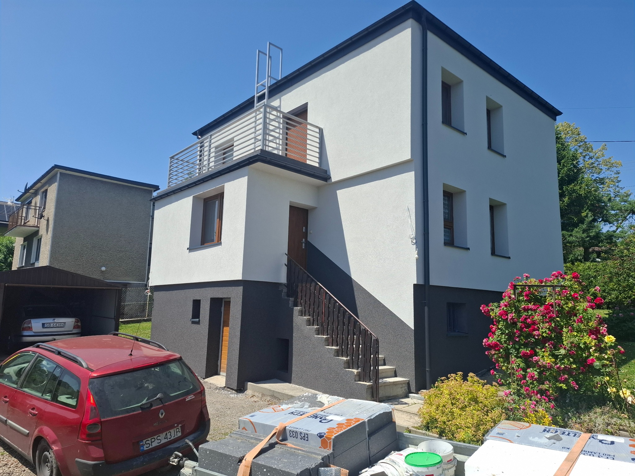 Dwukondygnacyjny dom po renowacji elewacji, z biało-szarą kolorystyką, balkonem i schodami wejściowymi. Na pierwszym planie materiały budowlane i samochód.