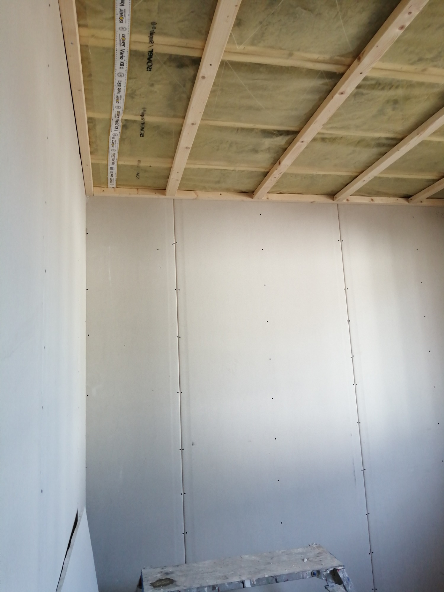 Wnętrze w trakcie montażu płyt kartonowo-gipsowych na ścianach i suficie, widoczna konstrukcja drewniana sufitu z izolacją, drabina malarska w rogu pomieszczenia.