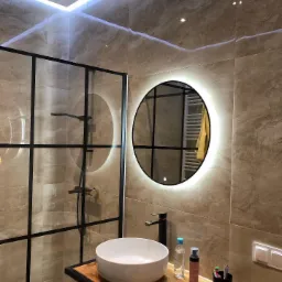 Nowoczesna łazienka z kabiną prysznicową typu walk-in z czarnymi szprosami, okrągłym lustrem z podświetleniem LED, umywalką nablatową na drewnianym blacie i marmurowymi płytkami na ścianach.