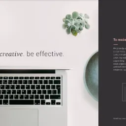 Laptop z tekstem 'be creative. be effective.' na białym biurku obok sukulentu w doniczce, świeczki i notatnika, widok z góry.
