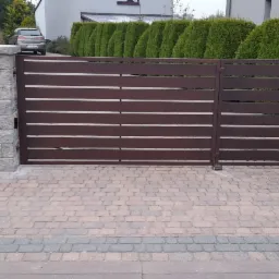 Nowoczesna brama wjazdowa z poziomych profili w kolorze brązowym, osadzona na słupkach z szarego kamienia łupanego, na tle kostki brukowej i zadbanego żywopłotu.