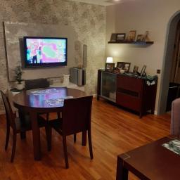 WITABUD WITOLD GOJ - Jasne wnętrze pokoju z drewnianą podłogą, okrągłym stołem i telewizorem na ścianie z wzorzystą tapetą.