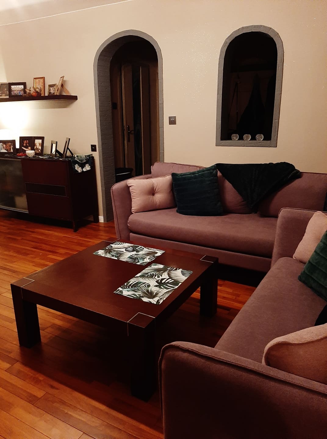 Salon z fioletową sofą, drewnianym stolikiem kawowym z podkładkami w liście i łukowatymi wnękami w ścianie, jedna z drzwiami, druga z dekoracjami.