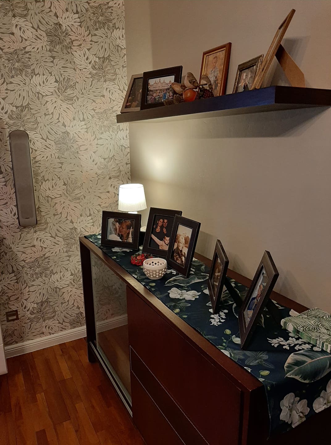 Pokój z wzorzystą tapetą w liście, półką z ramkami i komodą z dekoracyjnym bieżnikiem w kwiaty.