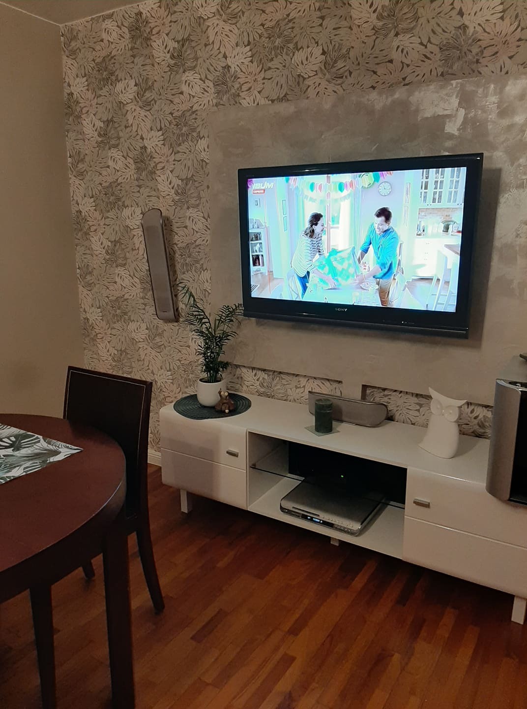 Salon z telewizorem LCD zawieszonym na ścianie z tapetą w liście, biała szafka RTV z dekoracjami, drewniany stół z krzesłem w rogu.