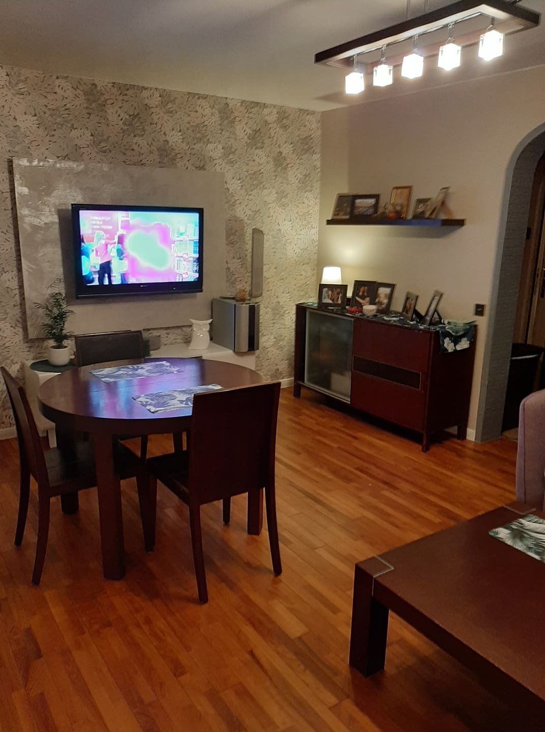 Jasne wnętrze pokoju z drewnianą podłogą, okrągłym stołem i telewizorem na ścianie z wzorzystą tapetą.