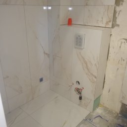 Firma Kornik Michał Modrak - Montaż stelaża podtynkowego WC z przyłączami wodnymi i płytkami imitującymi marmur na ścianach i podłodze, widoczne narzędzia i resztki kleju.