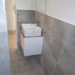 Firma Kornik Michał Modrak - Nowoczesna łazienka w trakcie wykańczania: biała szafka z umywalką na drewnianym blacie, szare płytki imitujące beton na ścianie, widoczny fragment lustra i niedokończone wykończenie przy drzwiach.