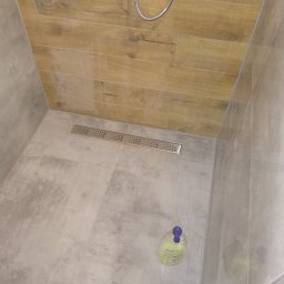 Firma Kornik Michał Modrak - Wykończony brodzik prysznicowy z odpływem liniowym, wyłożony płytkami imitującymi drewno na ścianach i szarymi płytkami na podłodze; butelka płynu do kąpieli w rogu.