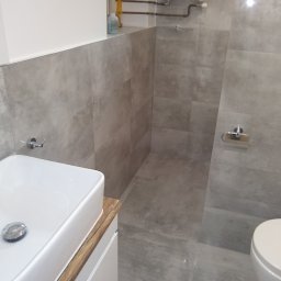 Firma Kornik Michał Modrak - Nowoczesna łazienka z umywalką nablatową na szafce z drewnianym blatem, szare płytki imitujące beton na ścianach i podłodze, oraz grzejnik drabinkowy.