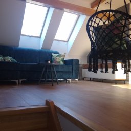 Firma Kornik Michał Modrak - Jasne poddasze z drewnianymi belkami, oknami dachowymi, granatową sofą z poduszkami w tropikalne wzory, okrągłym stolikiem kawowym i wiszącym fotelem brazylijskim z czarnego sznurka.