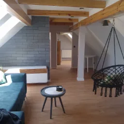 Jasne poddasze z drewnianymi belkami, szarą cegłą na ścianie, turkusową sofą i wiszącym fotelem hamakowym