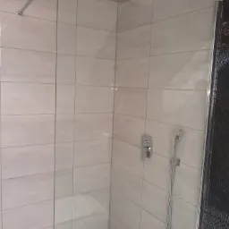 Nowoczesna kabina prysznicowa z deszczownicą, wykończona jasnymi, prostokątnymi płytkami i czarną mozaiką po prawej stronie, widoczny odpływ liniowy w podłodze.