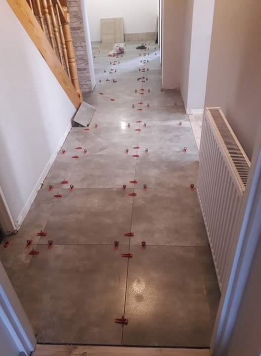 Układanie szarych płytek podłogowych w korytarzu, widoczne czerwone klipsy poziomujące, schody z drewnianą balustradą w tle.