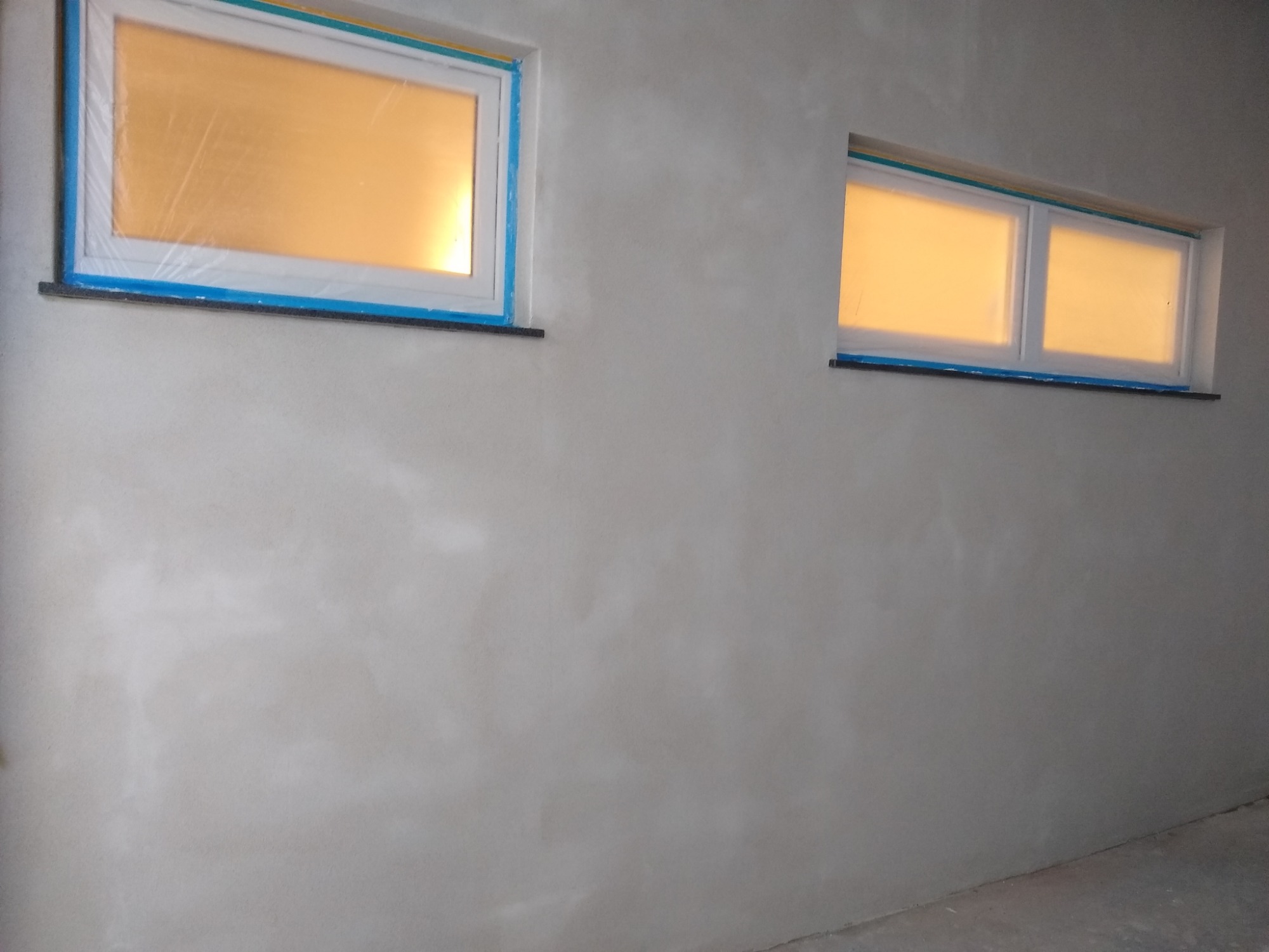 Świeżo otynkowana szara ściana wewnętrzna z dwoma oknami zabezpieczonymi folią i niebieską taśmą malarską.