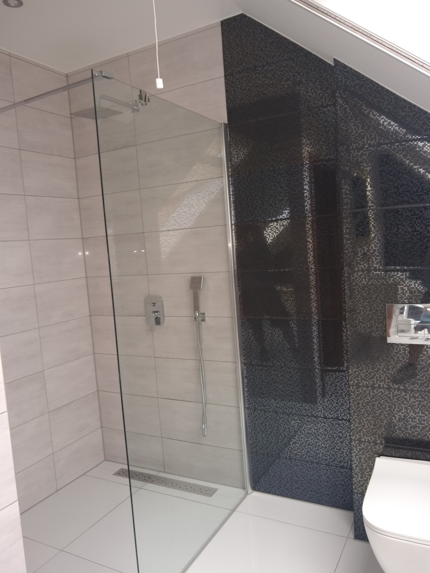 Nowoczesna kabina prysznicowa z deszczownicą i szklanymi drzwiami, wykończona białymi płytkami i mozaiką w odcieniach szarości i czerni, widoczny odpływ liniowy w podłodze.