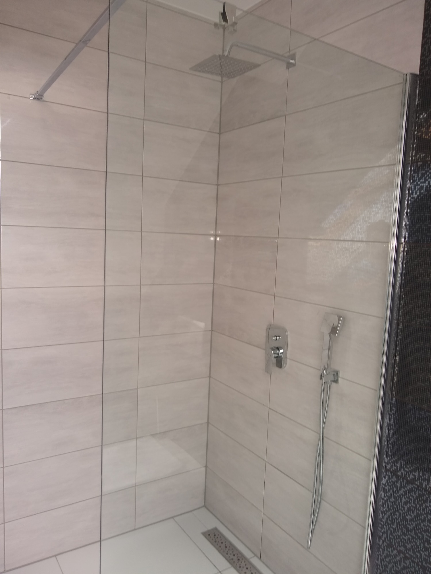 Nowoczesna kabina prysznicowa z deszczownicą, wykończona jasnymi, prostokątnymi płytkami i czarną mozaiką po prawej stronie, widoczny odpływ liniowy w podłodze.