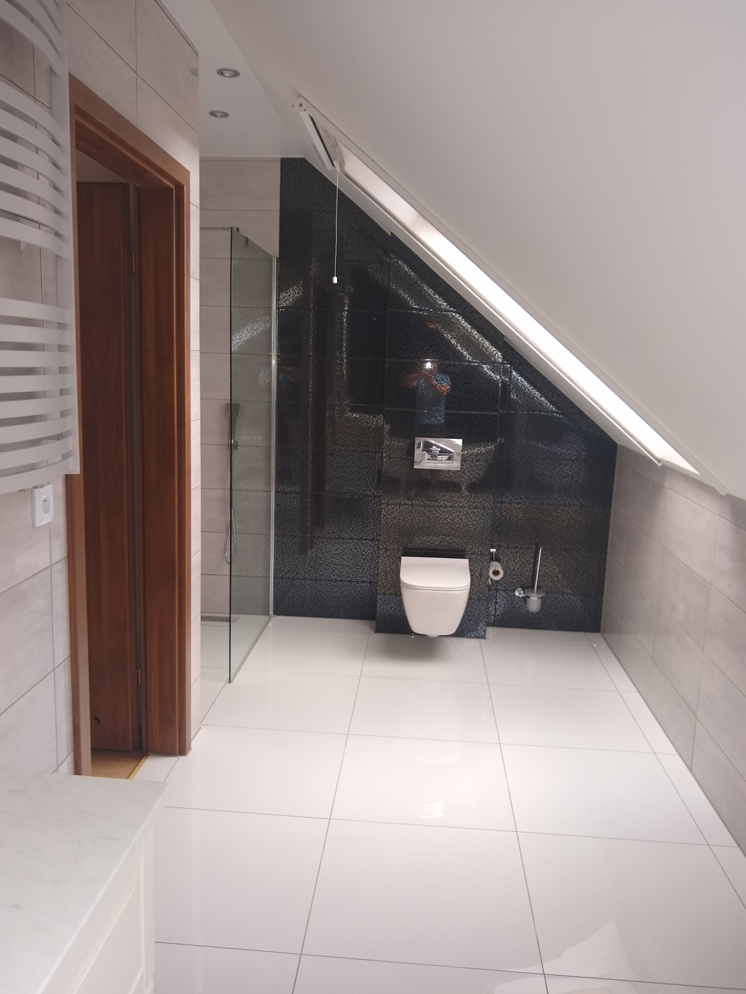 Nowoczesna łazienka na poddaszu z białą armaturą, czarną mozaiką na ścianie ze skosem i białymi płytkami podłogowymi z ciemną fugą.