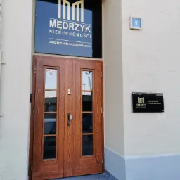 Wejście do biura nieruchomości Mędrzyk z drewnianymi drzwiami i tablicami informacyjnymi na elewacji.