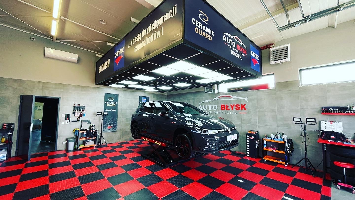 Samochód na podnośniku w warsztacie detailingowym Auto Błysk, widoczne logo Ceramic Guard, podłoga w czarno-czerwoną szachownicę, profesjonalne oświetlenie i wyposażenie.
