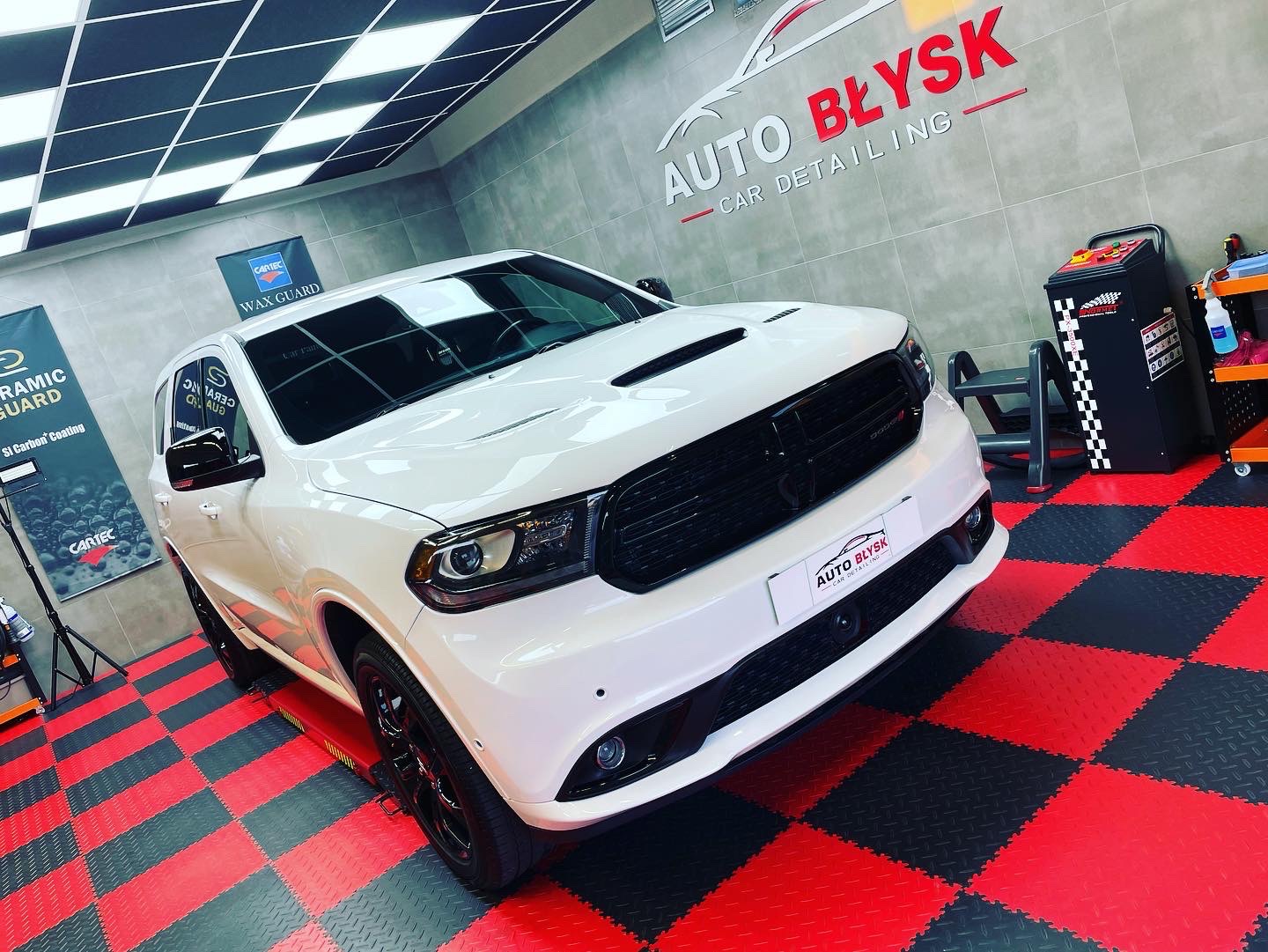Biały Dodge Durango po detailingu, z logo 'Auto Błysk' w tle, na czerwono-czarnej podłodze w kratkę, pod kątem.