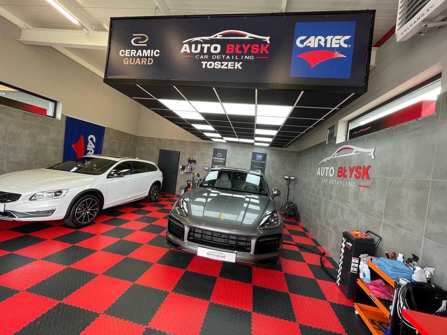 Wnętrze studia auto detailingu z dwoma samochodami (biały SUV i szary Porsche) na podłodze w czerwoną i czarną szachownicę, widoczne logo firmy Auto Błysk i banery reklamowe Ceramic Guard oraz Cartec.