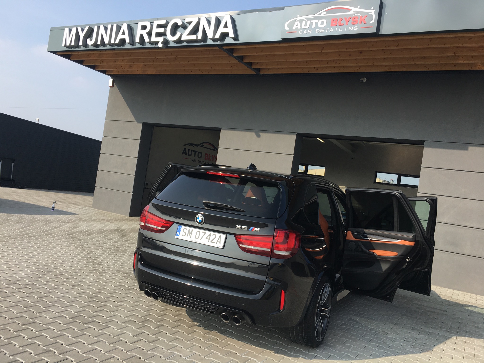 Czarny SUV BMW X5 M z otwartymi drzwiami na zewnątrz myjni ręcznej Auto Błysk, widok z tyłu, tablica rejestracyjna SM 0742A.