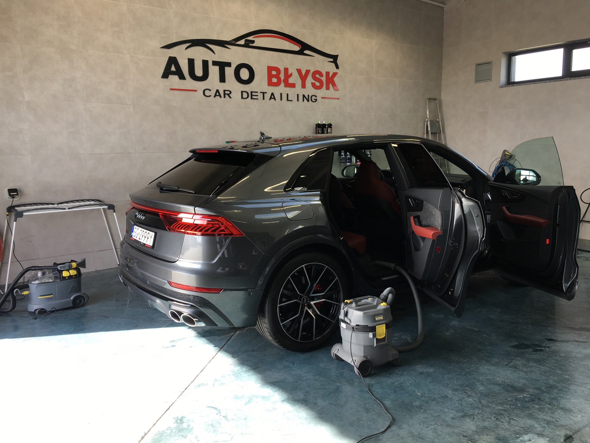 Szary SUV z otwartymi drzwiami wewnątrz myjni samochodowej, widoczny odkurzacz piorący Karcher i logo firmy Auto Błysk Car Detailing na ścianie.