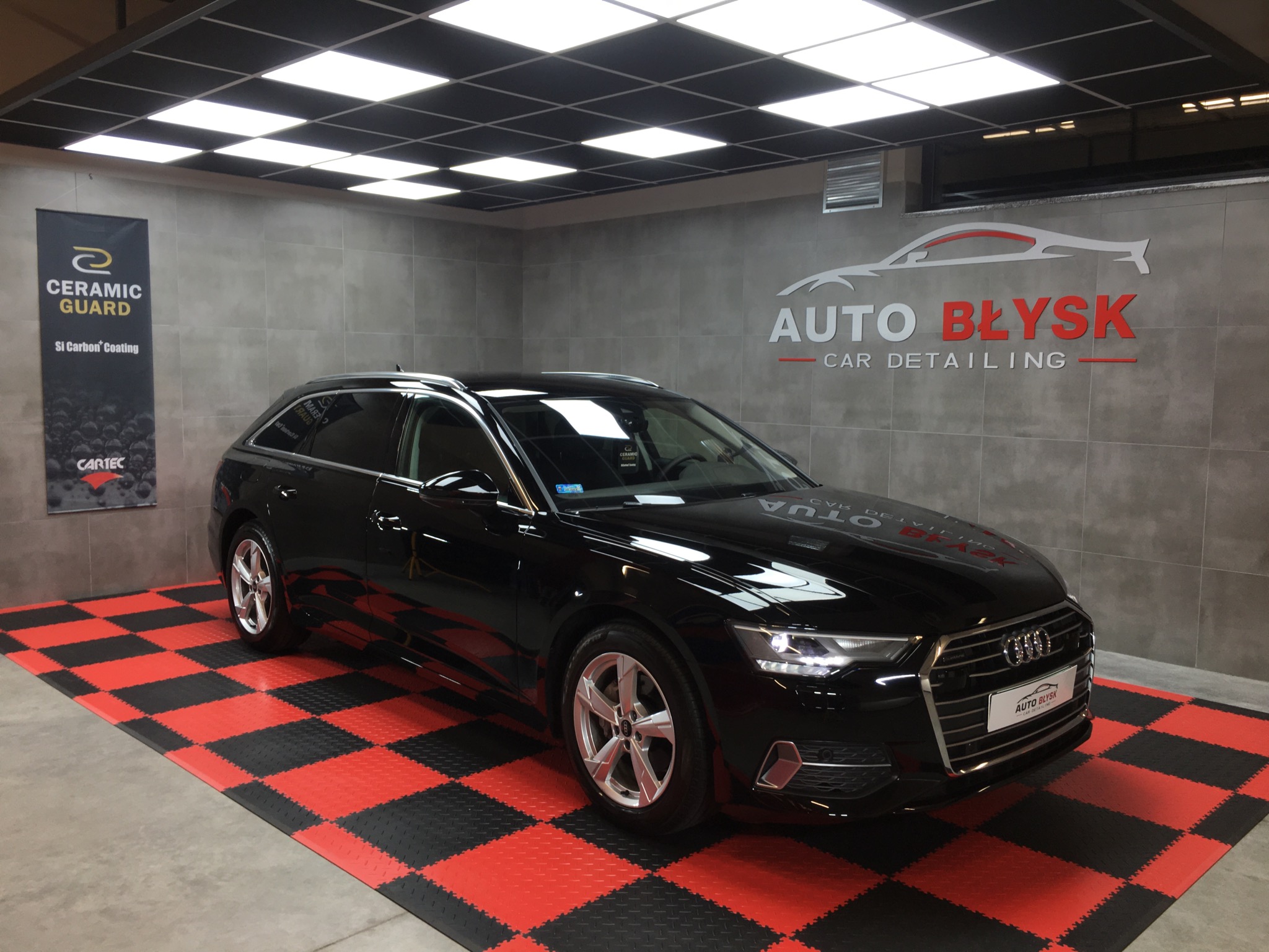 Błyszczący, czarny samochód kombi Audi po nałożeniu powłoki ceramicznej, zaparkowany w studiu detailingowym z logo 'Auto Błysk' na ścianie i banerem 'Ceramic Guard'.