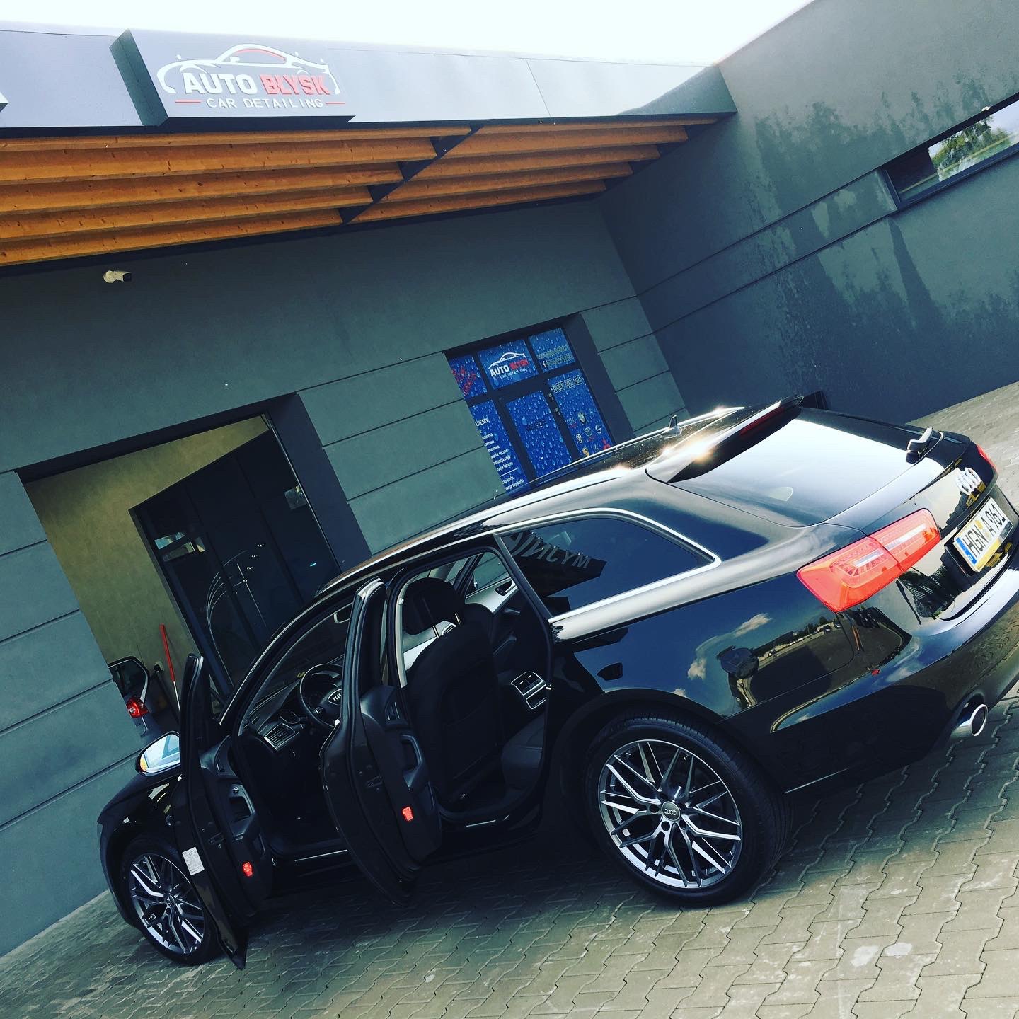 Czarny samochód kombi marki Audi z otwartymi drzwiami zaparkowany przed budynkiem myjni Auto Błysk Car Detailing z widocznym logo na elewacji i niebieskimi plakatami w oknach.