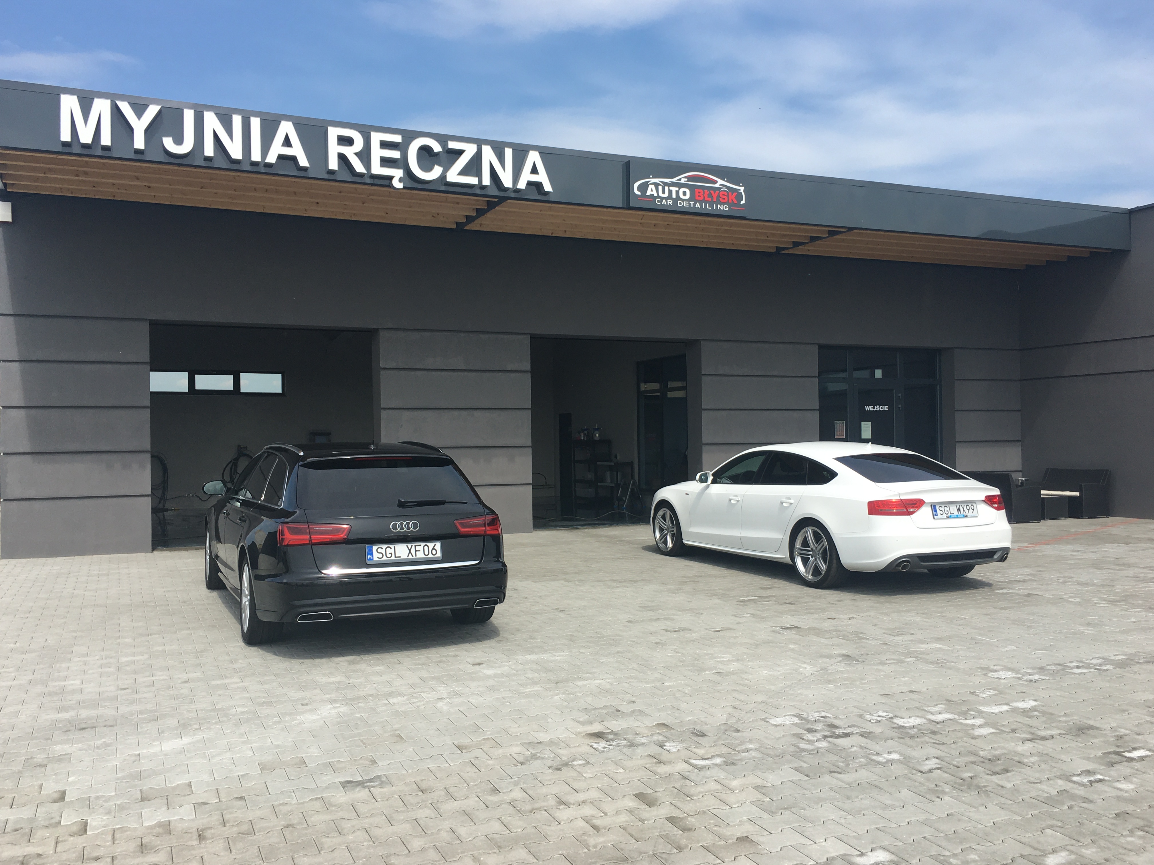 Czarny i biały samochód zaparkowane przed budynkiem myjni ręcznej z logo Auto Błysk Car Detailing w słoneczny dzień.