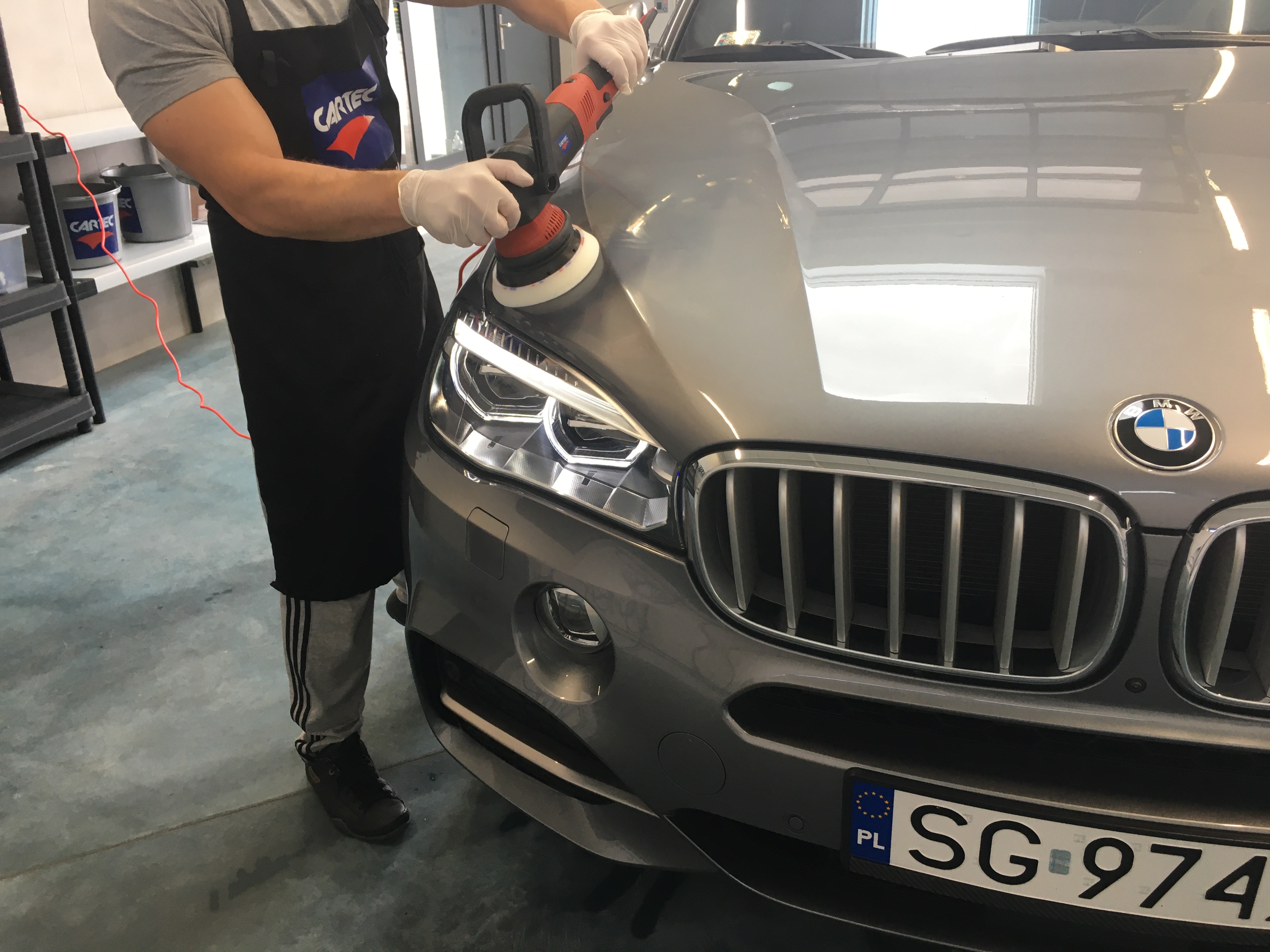 Pracownik w czarnym fartuchu poleruje przedni reflektor szarego samochodu BMW za pomocą elektrycznej polerki, widoczny fragment tablicy rejestracyjnej.