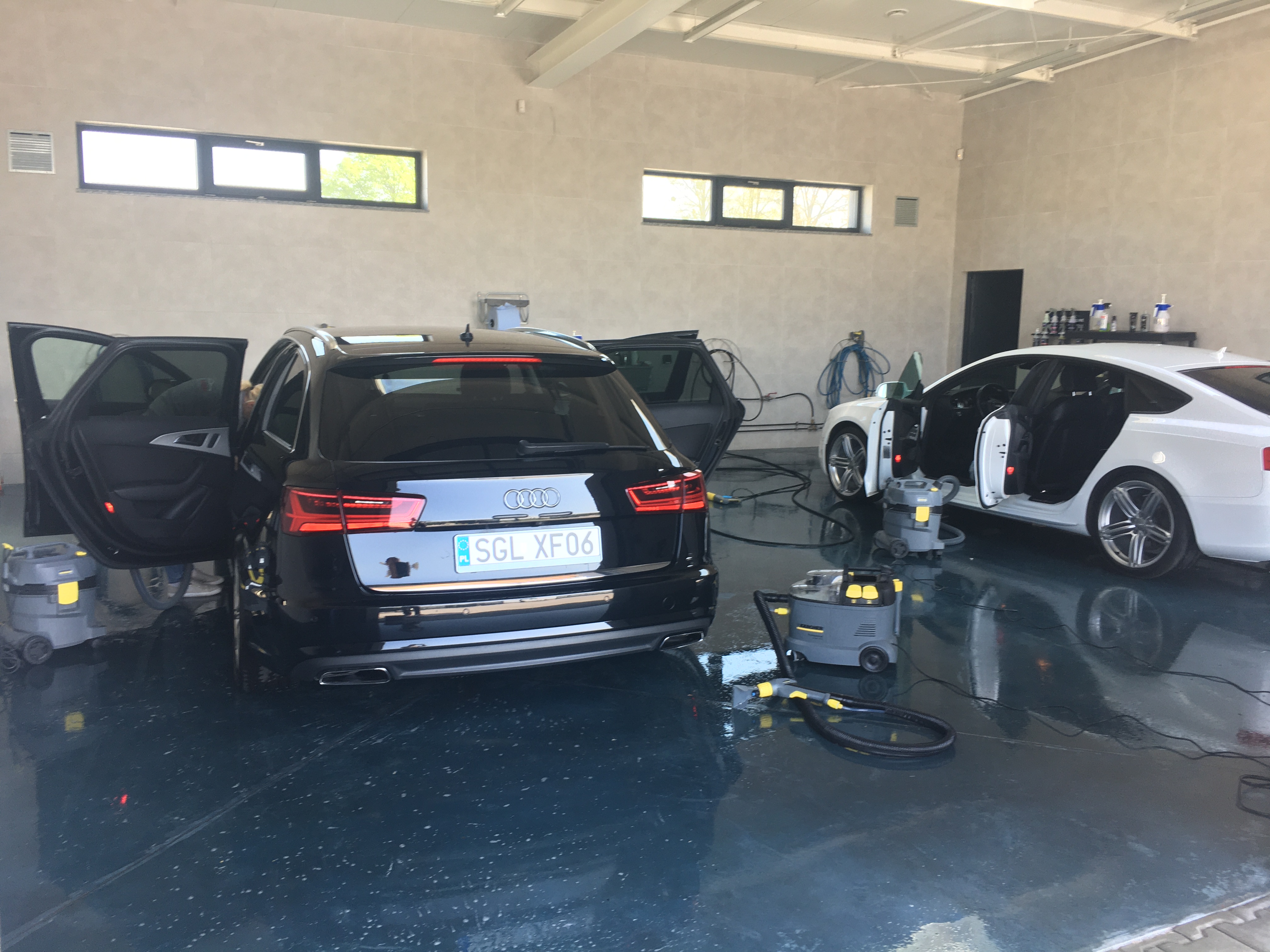 Dwa samochody marki Audi, czarny kombi i biały sedan, z otwartymi drzwiami w jasnym pomieszczeniu myjni samochodowej; widoczne odkurzacze piorące marki Karcher na mokrej, błyszczącej posadzce.