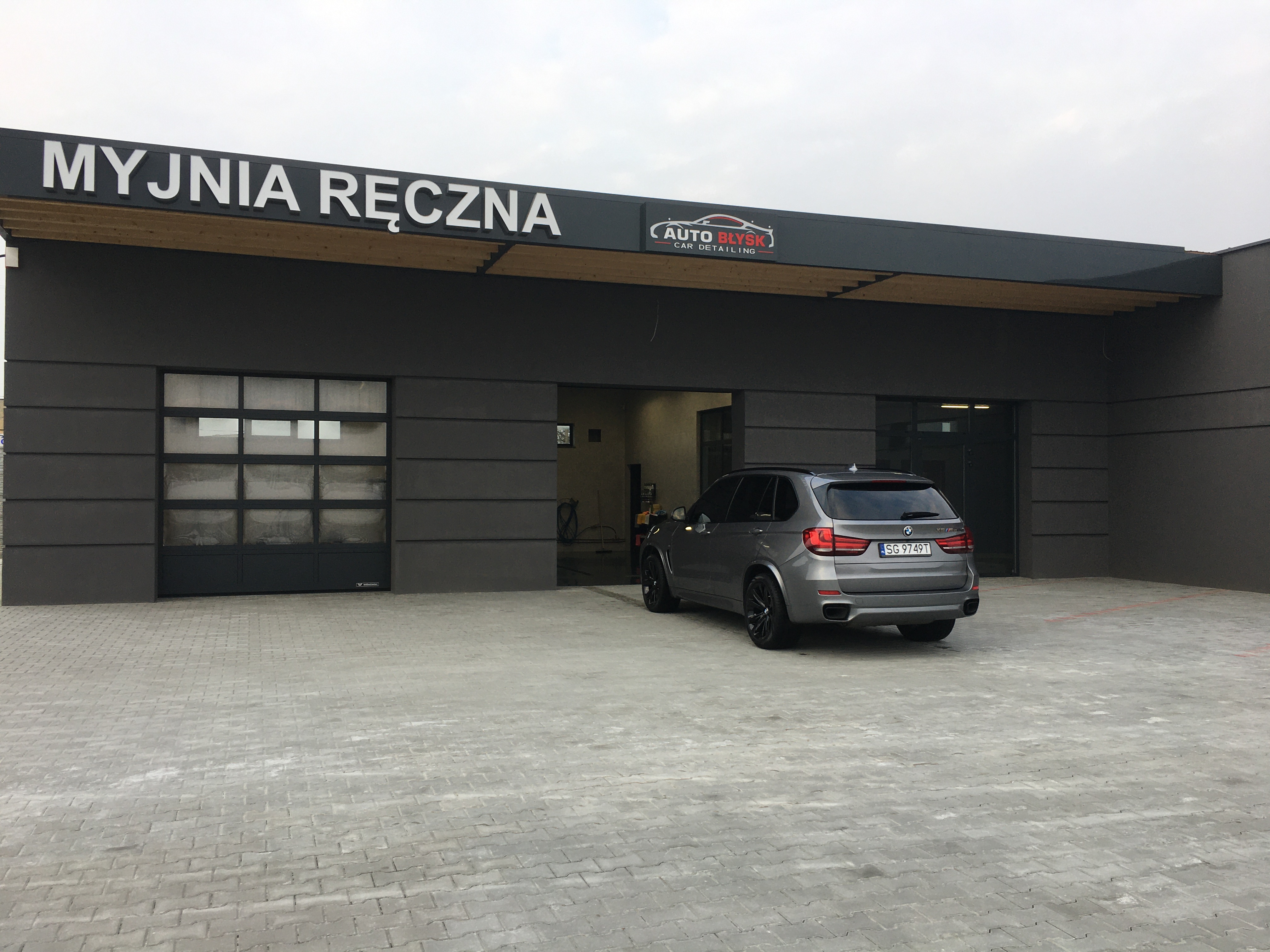 Szary samochód BMW X5 zaparkowany przed nowoczesną myjnią ręczną z szarym budynkiem i dużym napisem 'MYJNIA RĘCZNA' oraz logotypem 'AUTO BŁYSK CAR DETAILING' nad wjazdem.