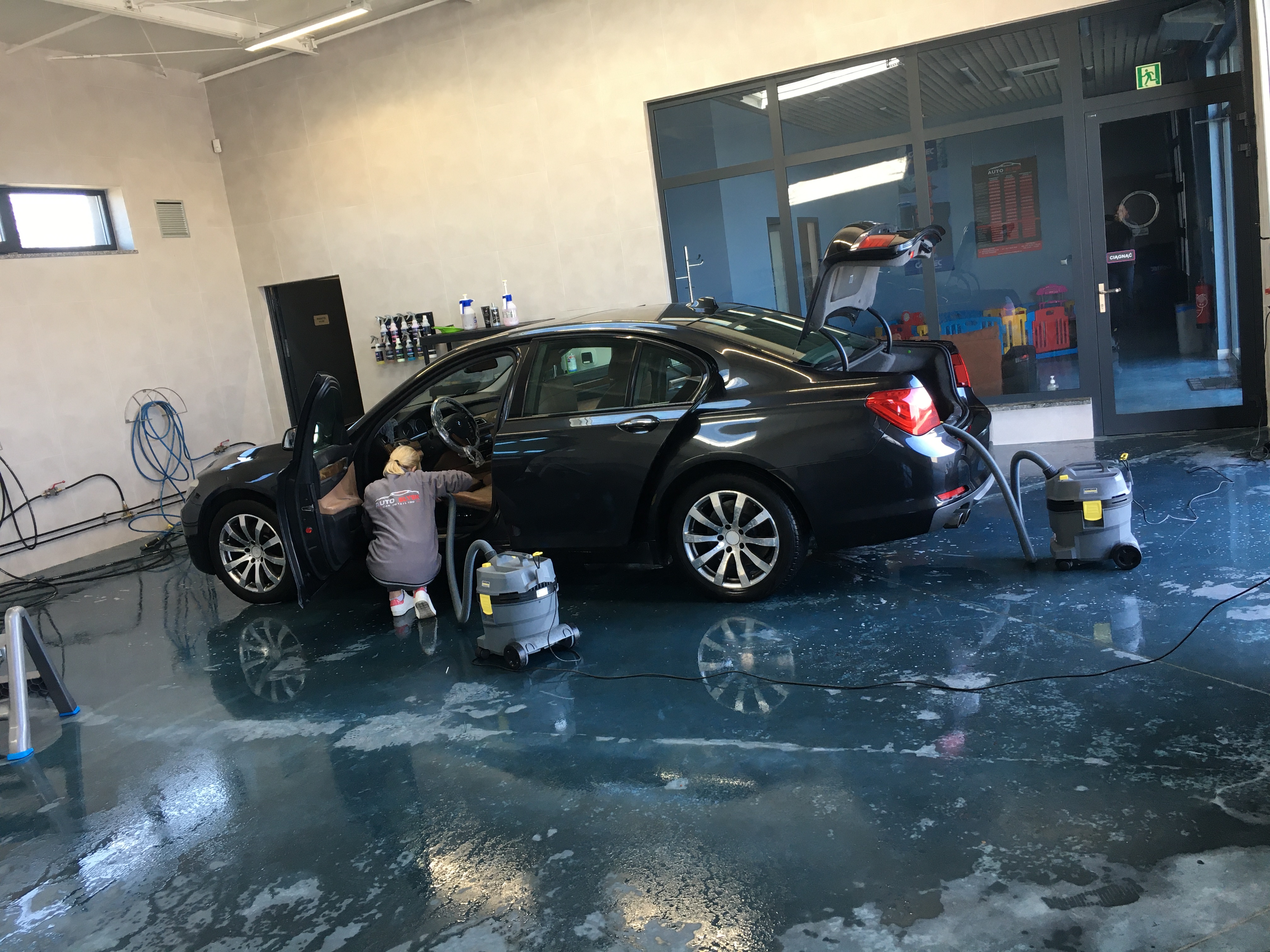 Kobieta w szarej bluzie z logo firmy czyści wnętrze ciemnego samochodu odkurzaczem przemysłowym w pomieszczeniu myjni samochodowej z mokrą podłogą.