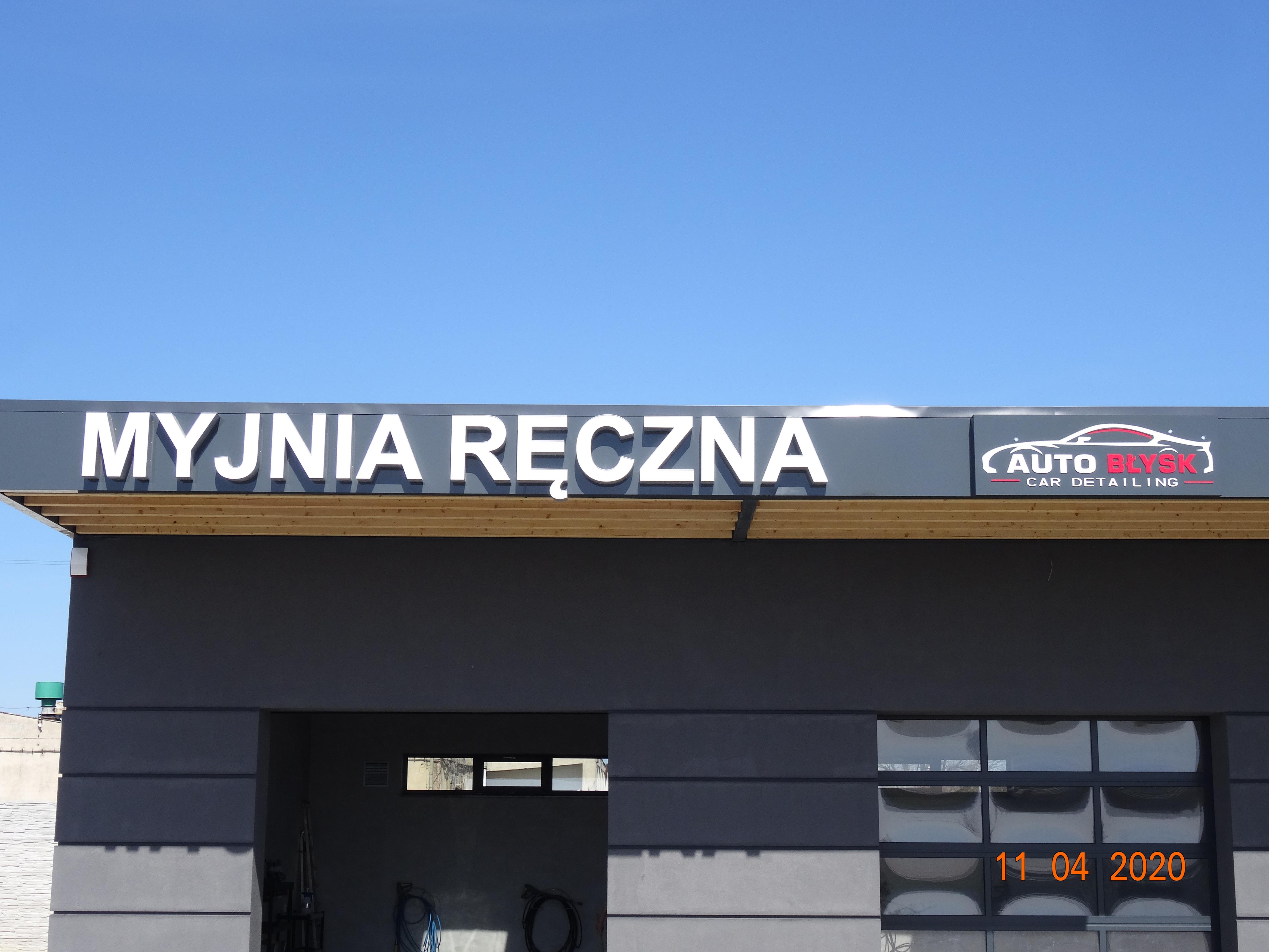 Elewacja budynku z szyldem 'Myjnia Ręczna' oraz logo 'Auto Błysk Car Detailing' na tle błękitnego nieba, widoczne wejście do myjni z narzędziami.